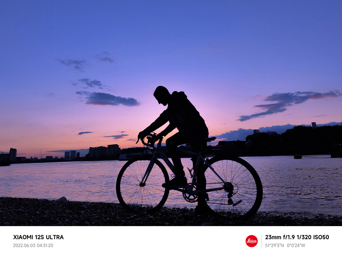Beispielaufnahme mit einem Xiaomi 12S Ultra, die einen Radfahrer am frühen Morgen vor Sonnenaufgang an einem Flussufer zeigt.